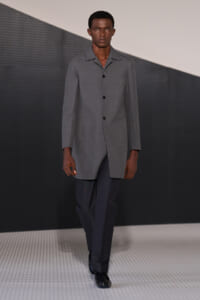 Male model walking the runway in a light gray, knee-length overcoat with black buttons and dark trousers, black shoes, on a geometric stage backdrop.