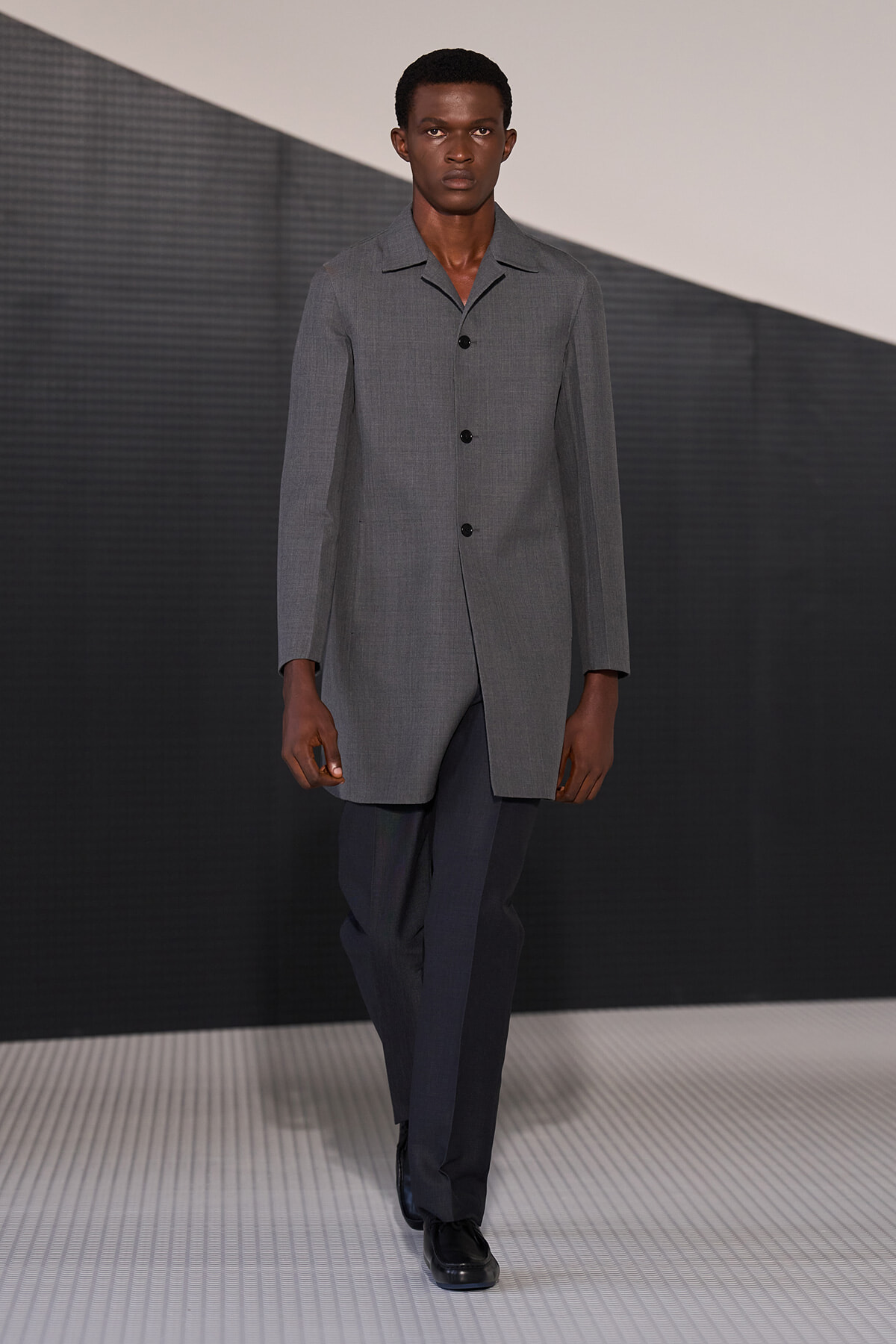 Male model walking the runway in a light gray, knee-length overcoat with black buttons and dark trousers, black shoes, on a geometric stage backdrop.