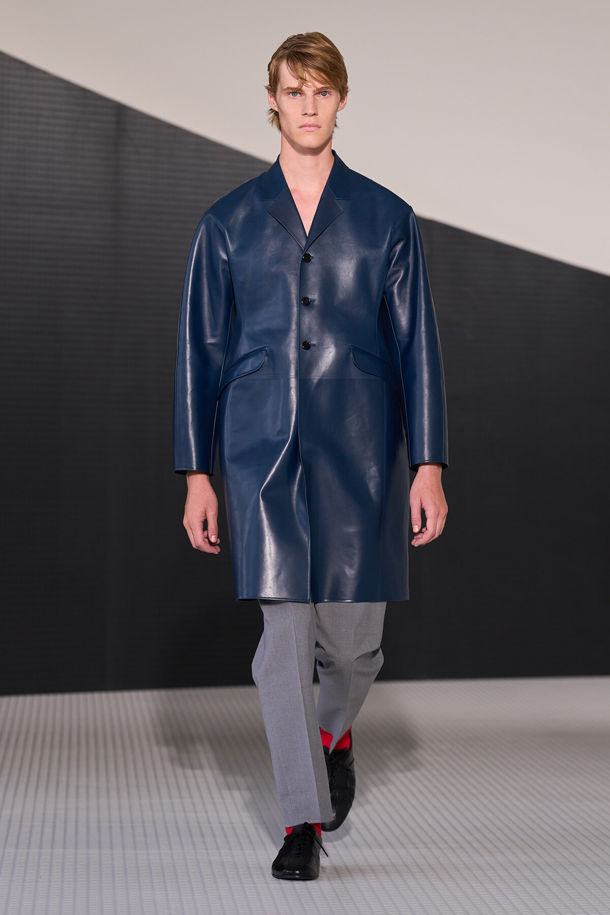 Male model on a runway wearing a navy blue knee-length leather coat, with gray trousers and black shoes, red socks showing at the ankles.