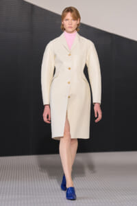 Model walking a fashion runway in a cream knee-length coat with gold buttons over a pink turtleneck, paired with blue shoes.