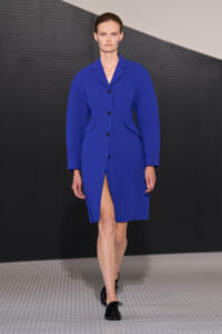 Model walks a cobalt blue knee-length coat with front buttons, side pocket flaps, and a front slit revealing her leg on a runway background.
