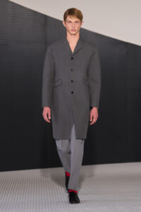 Male model walks a fashion runway wearing a gray knee-length overcoat with black buttons, paired with light gray trousers and black shoes with red socks showing at the ankles.