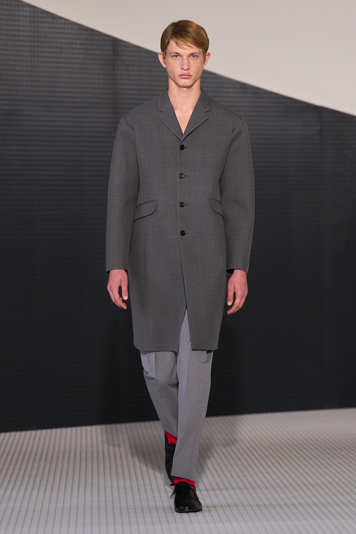 Male model walks a fashion runway wearing a gray knee-length overcoat with black buttons, paired with light gray trousers and black shoes with red socks showing at the ankles.