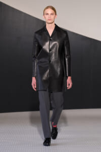 Male runway model wearing a black leather knee-length coat over gray trousers on a fashion catwalk.