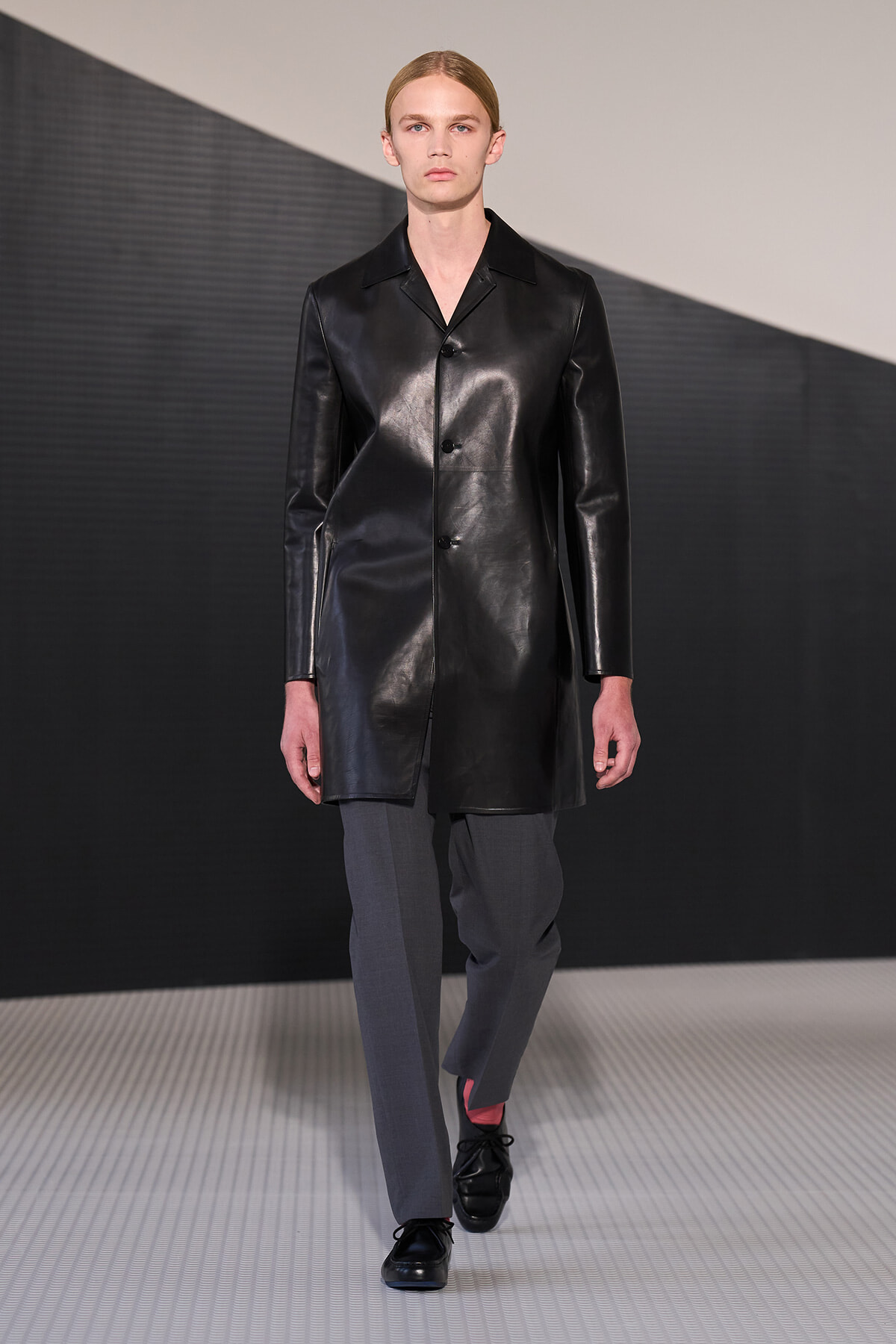 Male runway model wearing a black leather knee-length coat over gray trousers on a fashion catwalk.