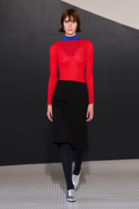 Model walking a runway in a red lace long-sleeve top over a blue base, paired with a black midi skirt, navy tights, and silver shoes.
