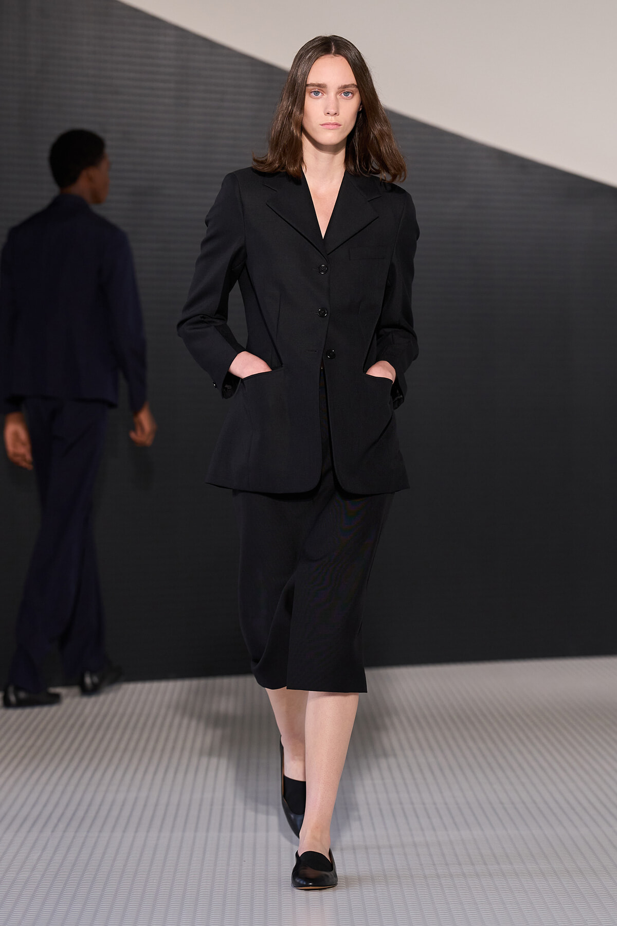 Female model in a black tailored blazer and knee-length black skirt walking the runway with hands in pockets.