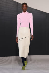 Model on a runway wearing a pastel pink turtleneck top and a cream knee-length skirt with a diagonal black swoosh, navy tights, and lime-green shoes.