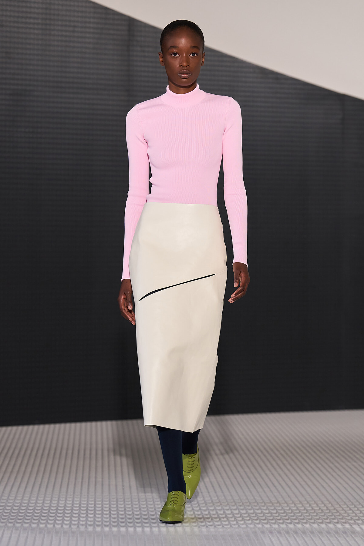 Model on a runway wearing a pastel pink turtleneck top and a cream knee-length skirt with a diagonal black swoosh, navy tights, and lime-green shoes.
