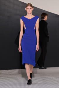 Model walking the runway in a cobalt blue, V-neck midi dress with side cutouts and black oxford shoes on a patterned floor.