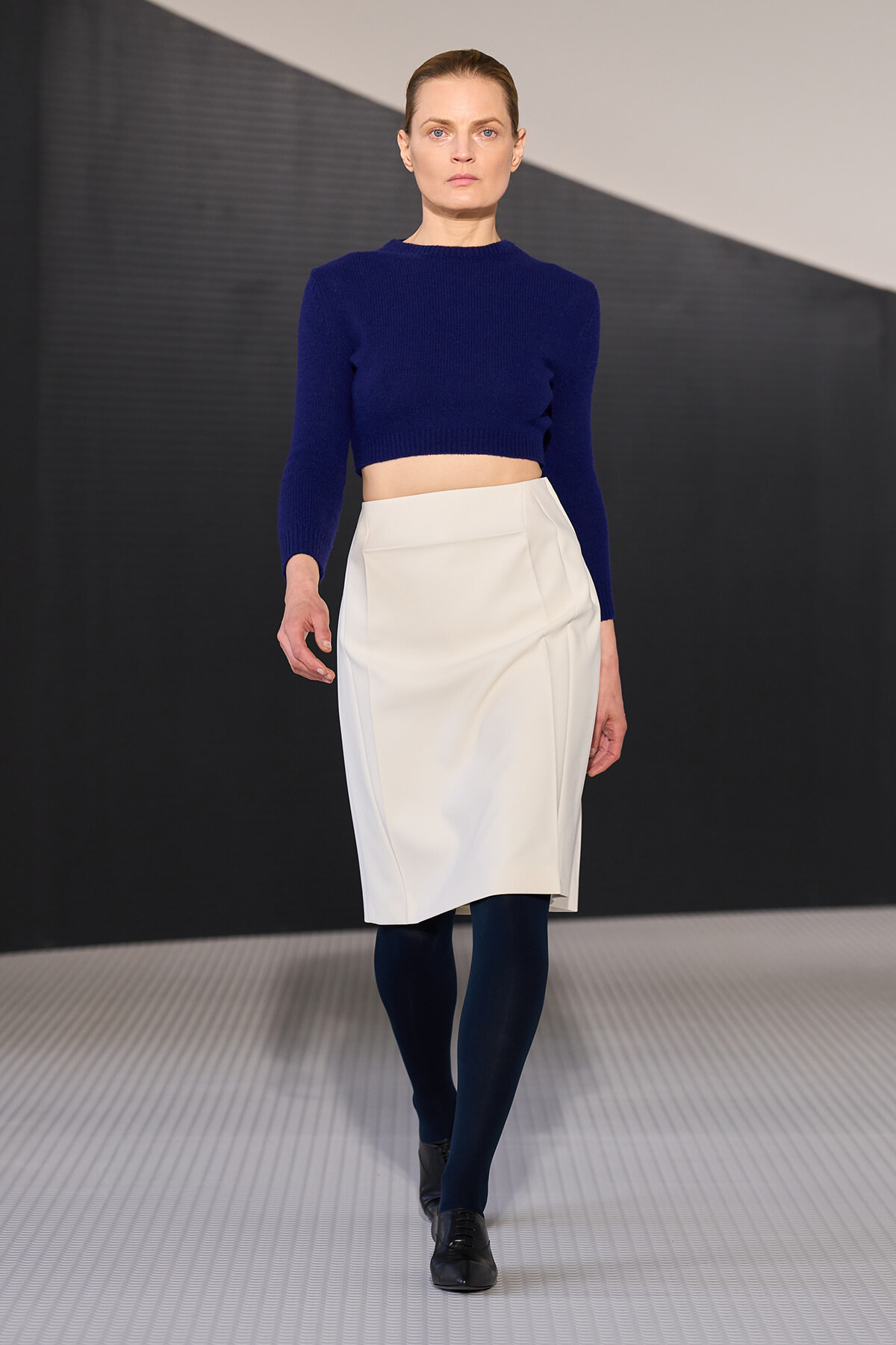 Model walking the runway in a royal blue cropped knit sweater and a white knee-length skirt, paired with navy tights and black shoes.