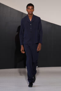 Male model walking a runway in a navy suit with a buttoned blazer and matching trousers.