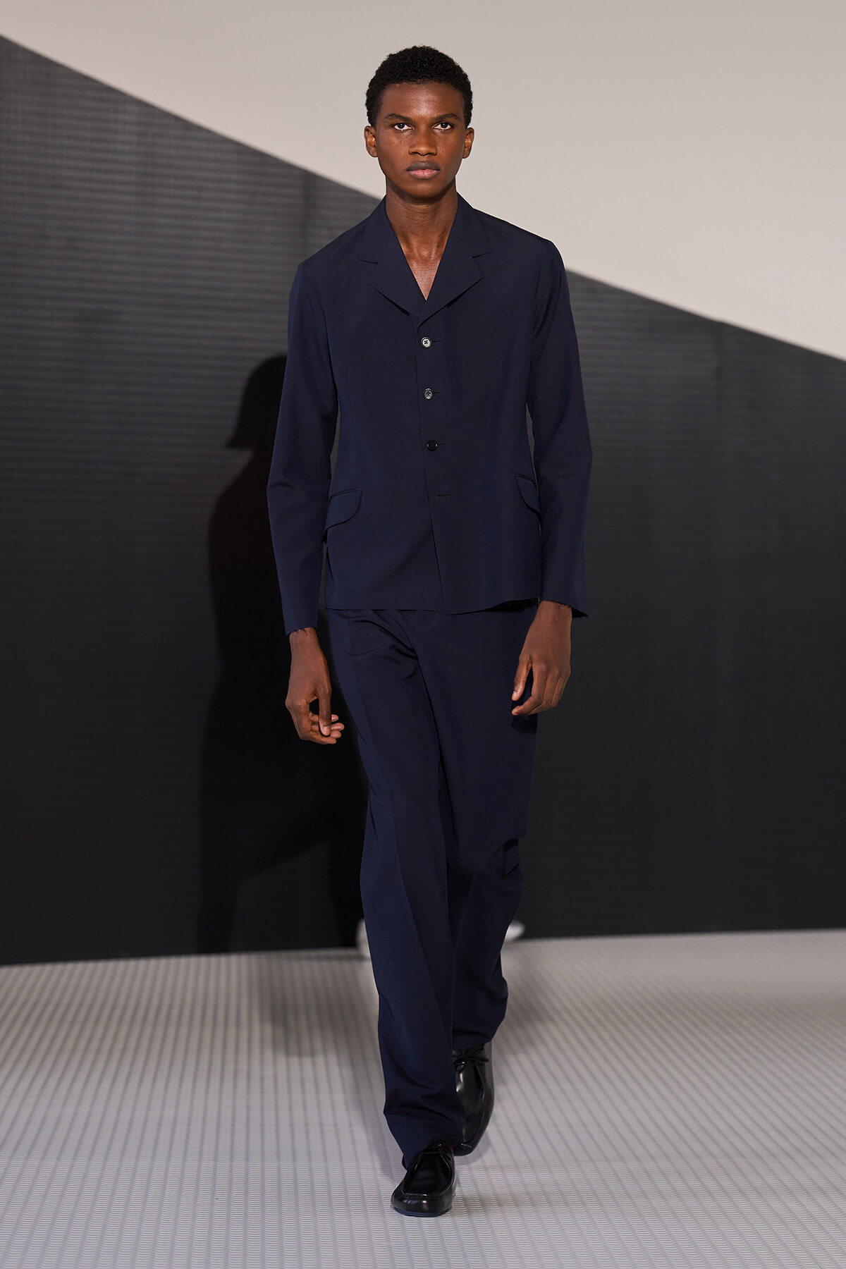 Male model walking a runway in a navy suit with a buttoned blazer and matching trousers.