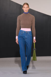 Model walking the runway in a brown turtleneck with pink cuffs and waistband, blue high-waisted jeans, black shoes, and a lime-green bag in hand.