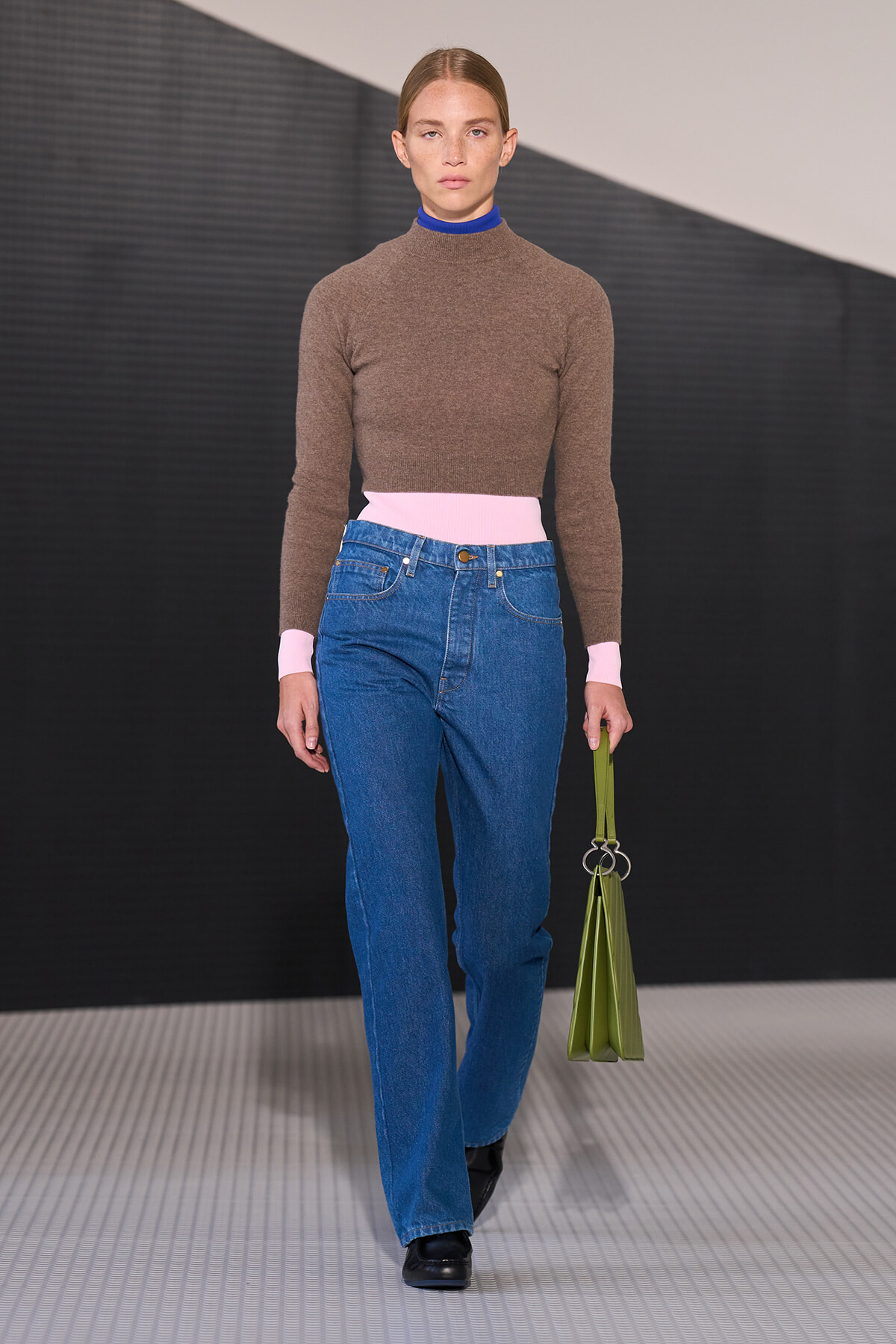 Model walking the runway in a brown turtleneck with pink cuffs and waistband, blue high-waisted jeans, black shoes, and a lime-green bag in hand.