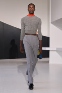 Male model walking a fashion runway in a grey cropped knit top, orange turtleneck, and light gray tailored trousers, black shoes