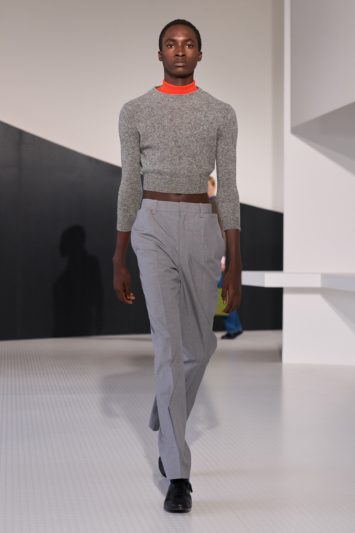 Male model walking a fashion runway in a grey cropped knit top, orange turtleneck, and light gray tailored trousers, black shoes