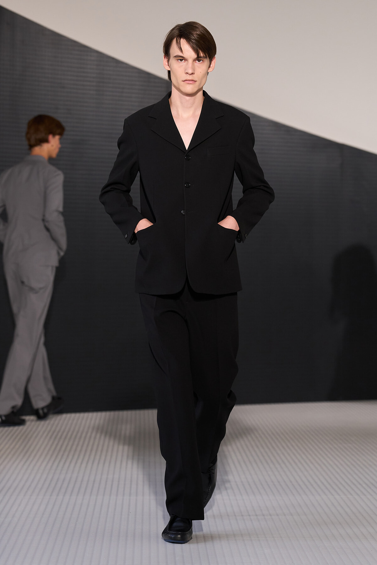 Male model in a black three‑button blazer and matching trousers walks the runway with hands in pockets.