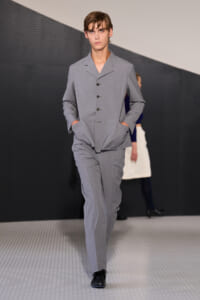Male model walks a fashion runway wearing a light gray tailored suit with jacket and matching pants, hands in pockets.