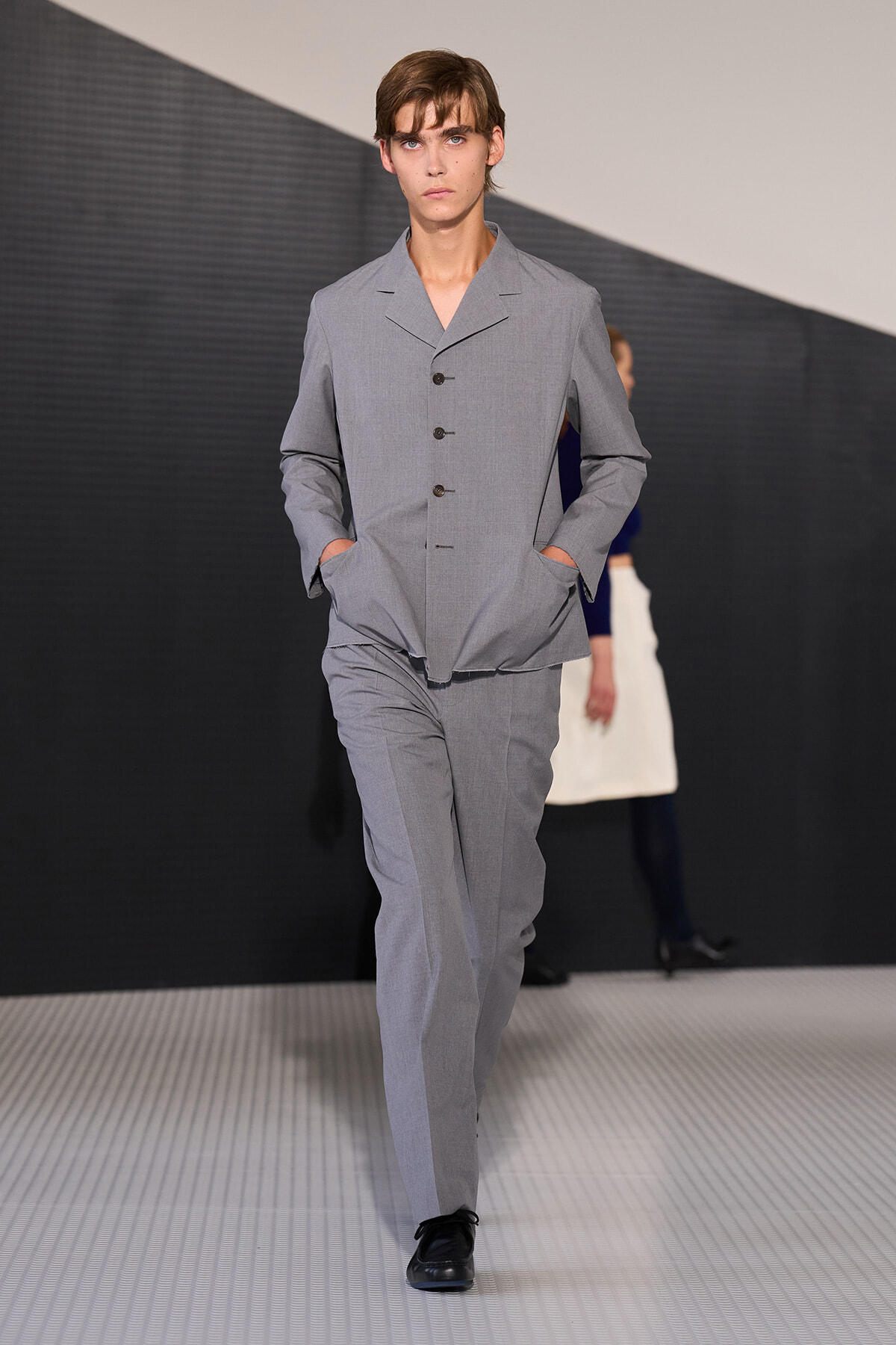 Male model walks a fashion runway wearing a light gray tailored suit with jacket and matching pants, hands in pockets.