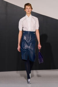 Runway model wearing a crisp white short-sleeve shirt with a navy leather midi skirt, navy tights, silver shoes, and a purple pleated handbag.
