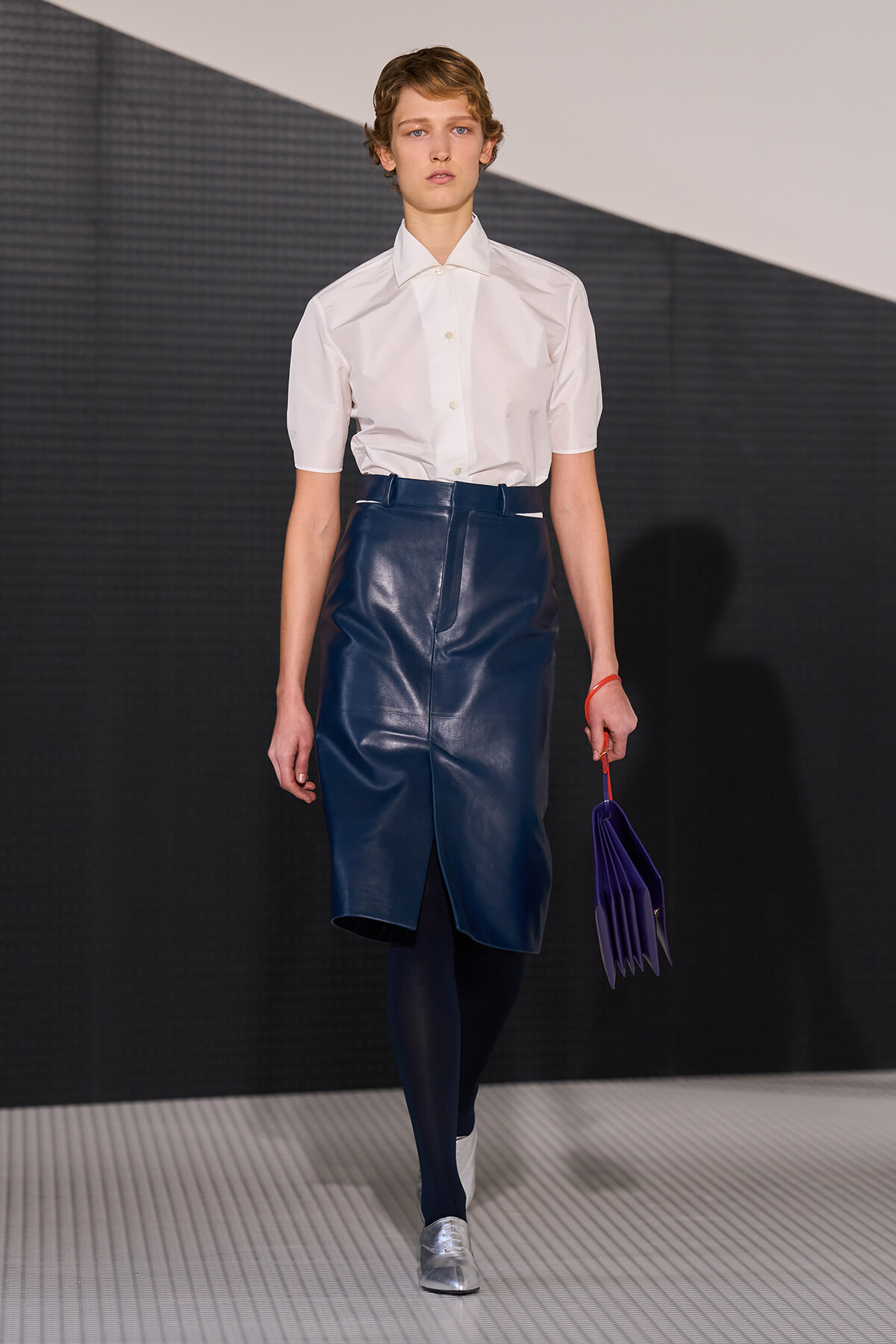 Runway model wearing a crisp white short-sleeve shirt with a navy leather midi skirt, navy tights, silver shoes, and a purple pleated handbag.