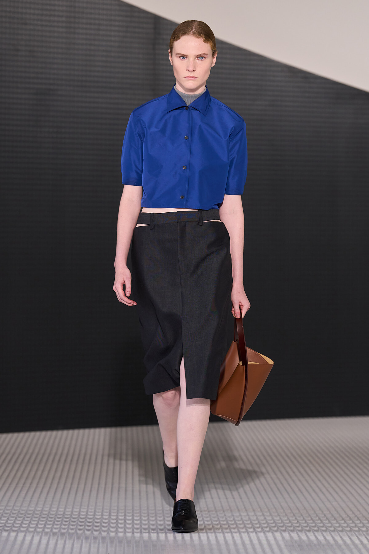Model wearing a cobalt cropped shirt with a dark midi skirt and brown tote on a runway.