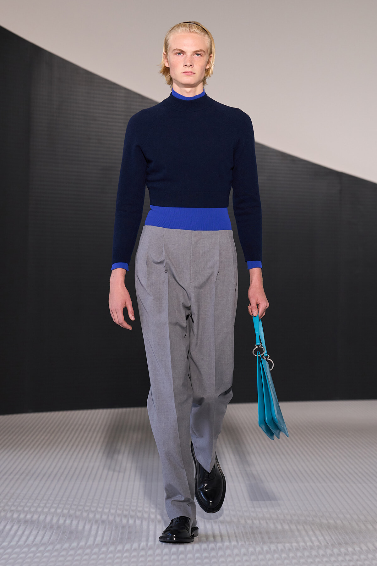 Male model on a runway wearing a navy sweater with a bright blue waistband and cuffs, light gray pleated trousers, black shoes, and carrying a turquoise handbag/portfolio.