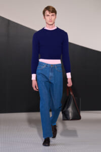 Male model on a runway wearing a navy knit sweater with pink cuffs and waistband, blue jeans, black shoes, and a black leather tote.
