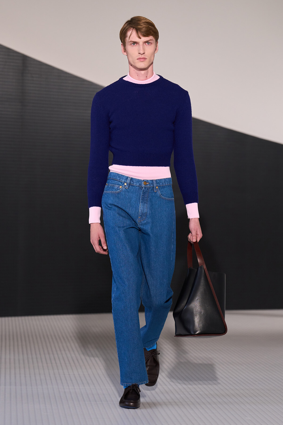 Male model on a runway wearing a navy knit sweater with pink cuffs and waistband, blue jeans, black shoes, and a black leather tote.