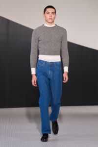 Male model walking a runway in a gray knit sweater with white cuffs and waistband, blue jeans, and black loafers.
