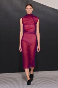 Model walks the runway in a sheer magenta sleeveless turtleneck dress with blue seam detailing, paired with black loafers against a dark backdrop.
