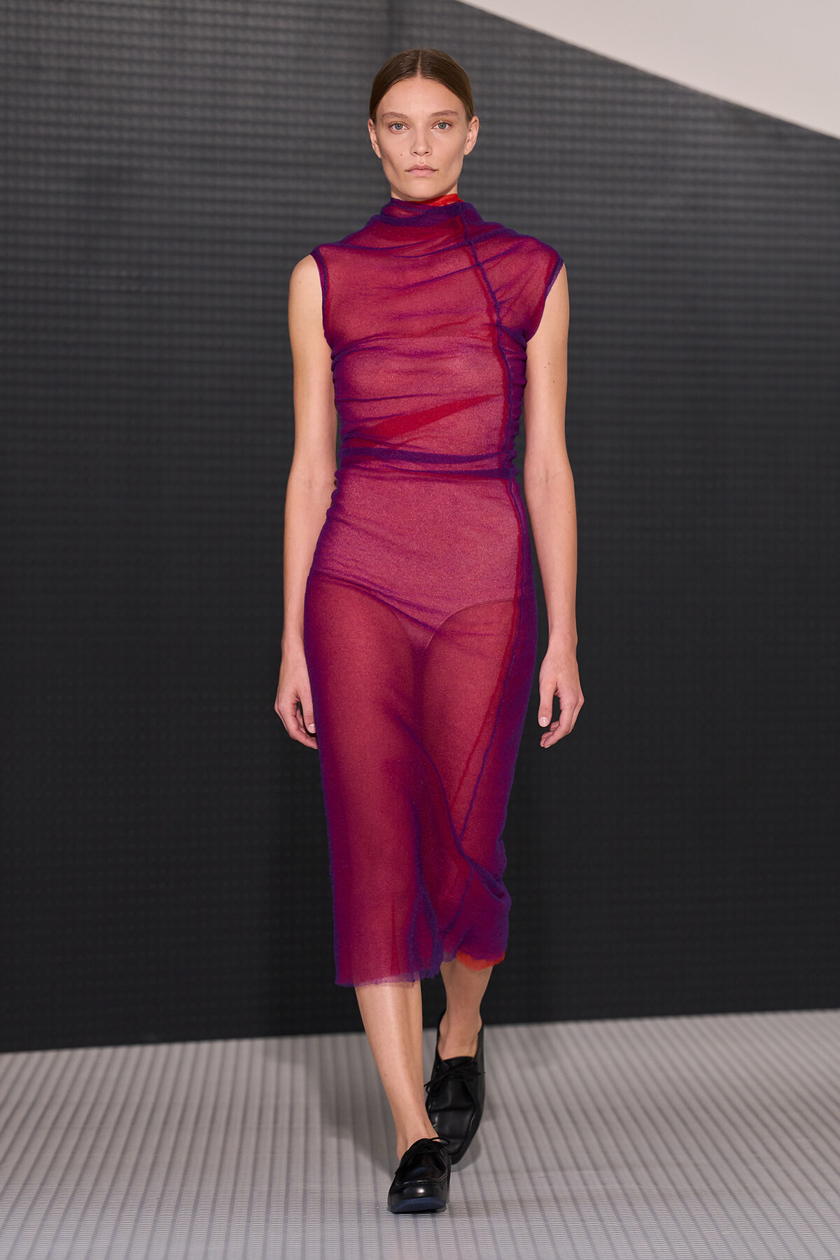 Model walks the runway in a sheer magenta sleeveless turtleneck dress with blue seam detailing, paired with black loafers against a dark backdrop.