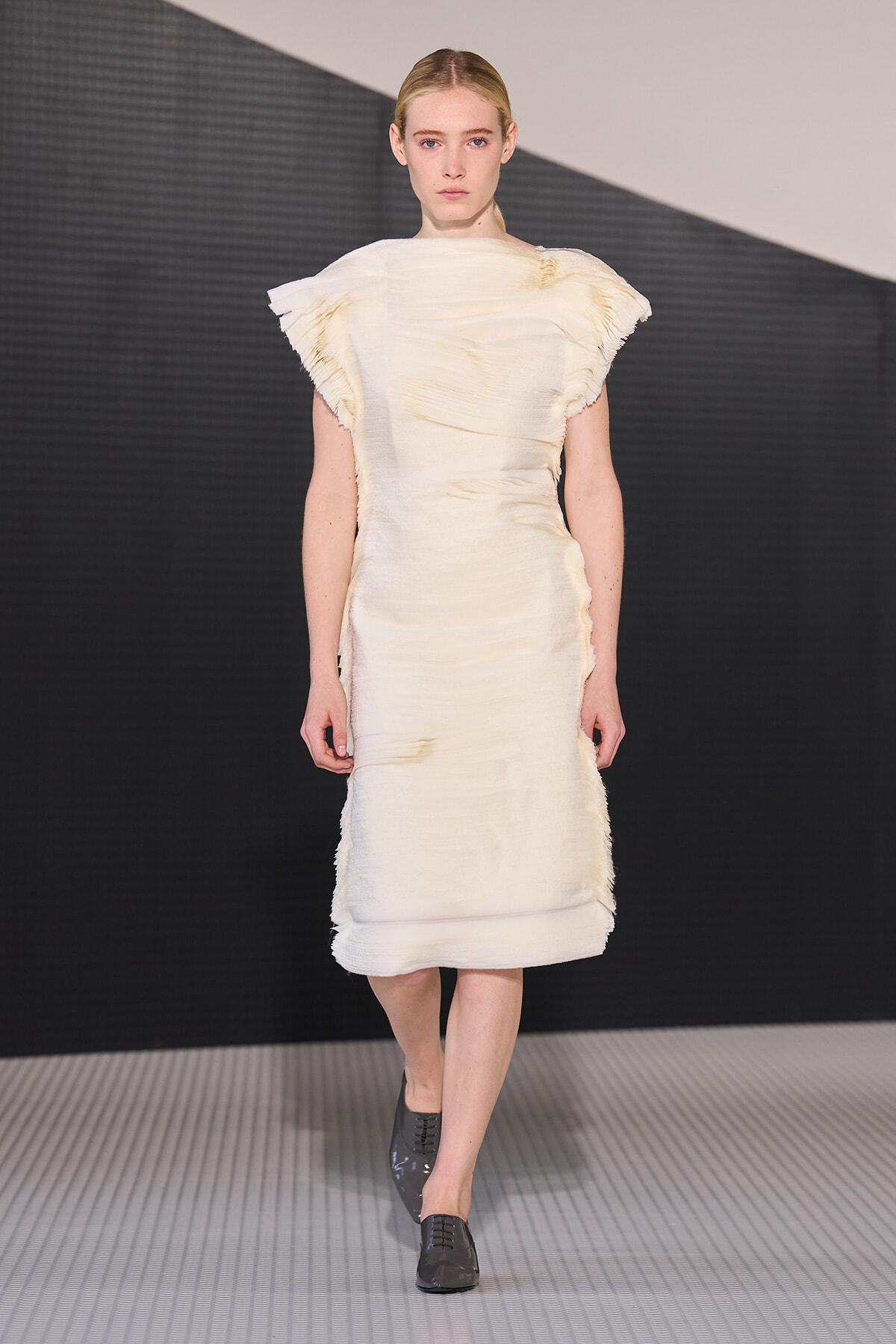 Model walking a runway in a pale cream, textured knee-length dress with sculpted, angular sleeves.