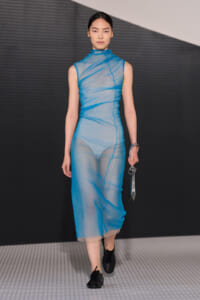 Model walking a fashion runway in a sheer blue sleeveless dress, holding a small metallic clutch.