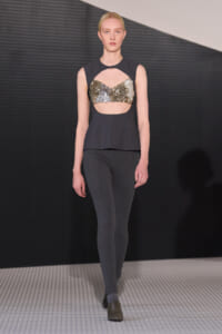 Model on a runway wearing a dark gray sleeveless top with a gold-sequin cutout bustier over a midriff, paired with slim gray pants.