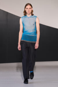 Male model walking a runway in a sheer blue sleeveless knit top and dark trousers, with black shoes, on a minimalist stage.