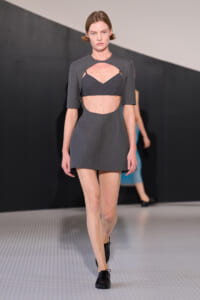 Female model walks the runway in a gray, structured mini dress with a cut-out midriff and a matching crop top beneath the bodice, black shoes.