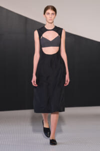 Model on a runway wearing a black sleeveless dress with a circular midriff cutout revealing a gray bralette underneath.