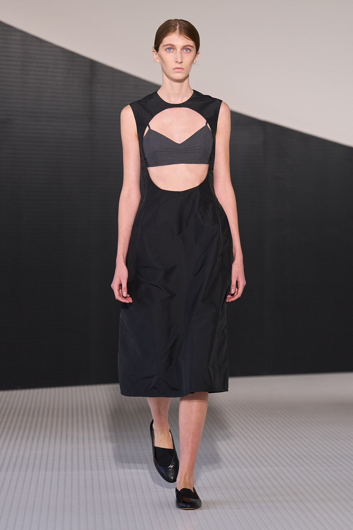 Model on a runway wearing a black sleeveless dress with a circular midriff cutout revealing a gray bralette underneath.