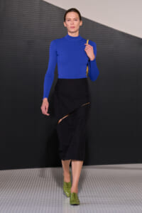 Model walking a fashion runway in a cobalt blue turtleneck, a black asymmetric wrap skirt with a side slit, lime-green oxford shoes, and a yellow bag over the shoulder.