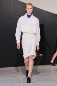 Model walking a fashion runway in a white cropped button-down jacket over a blue top paired with a knee-length white skirt, black shoes, and a large black-and-brown tote bag on her right arm.