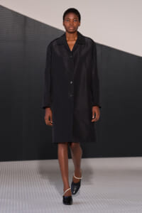 Model walks the runway in a knee-length black buttoned coat and black shoes with white straps, against a geometric backdrop.