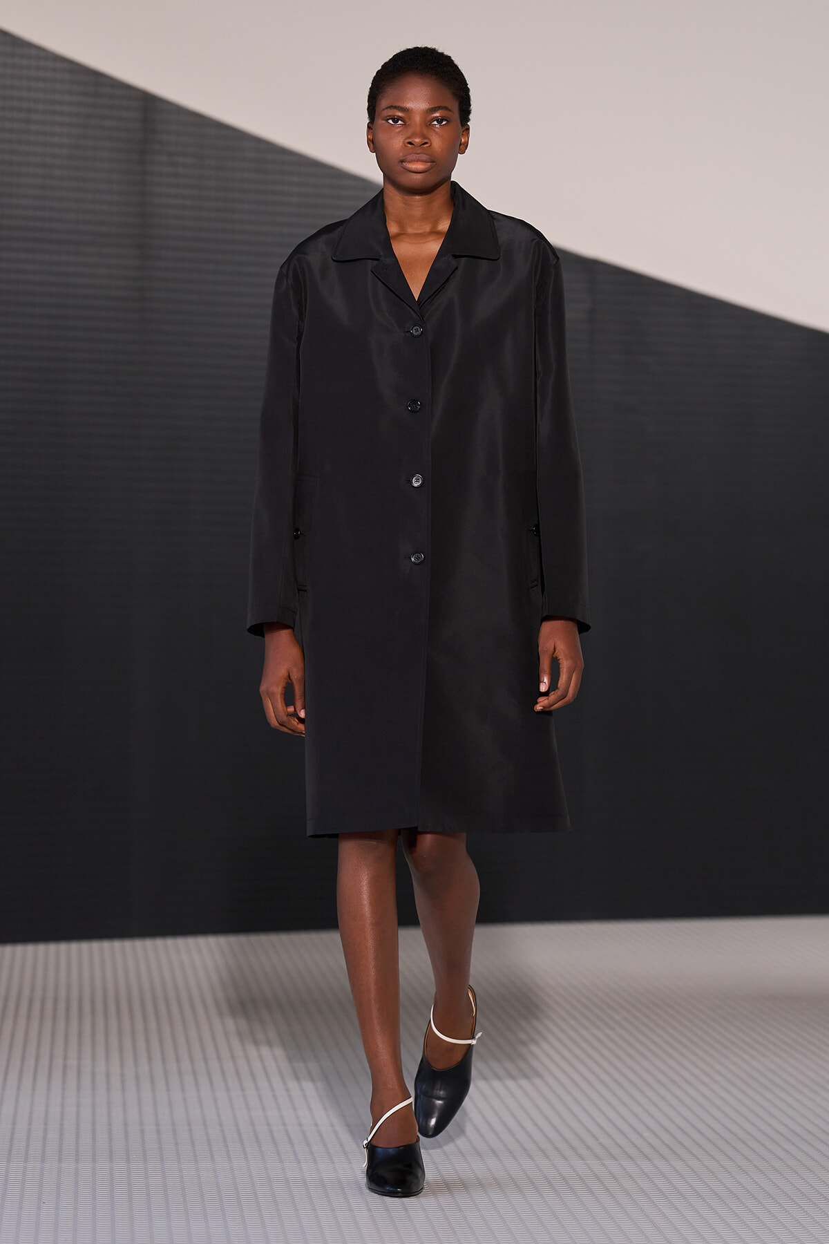 Model walks the runway in a knee-length black buttoned coat and black shoes with white straps, against a geometric backdrop.