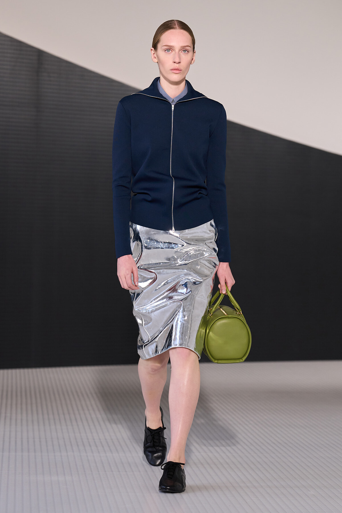 Model walking a runway in a navy zip cardigan over a light blue shirt, a metallic silver knee-length skirt, black shoes, and a lime green round handbag.