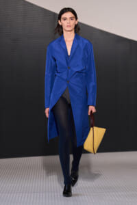 Model walks the runway in a bright blue overcoat-style blazer with an asymmetric wrap, black tights, black shiny shoes, and a yellow tote with brown handles.