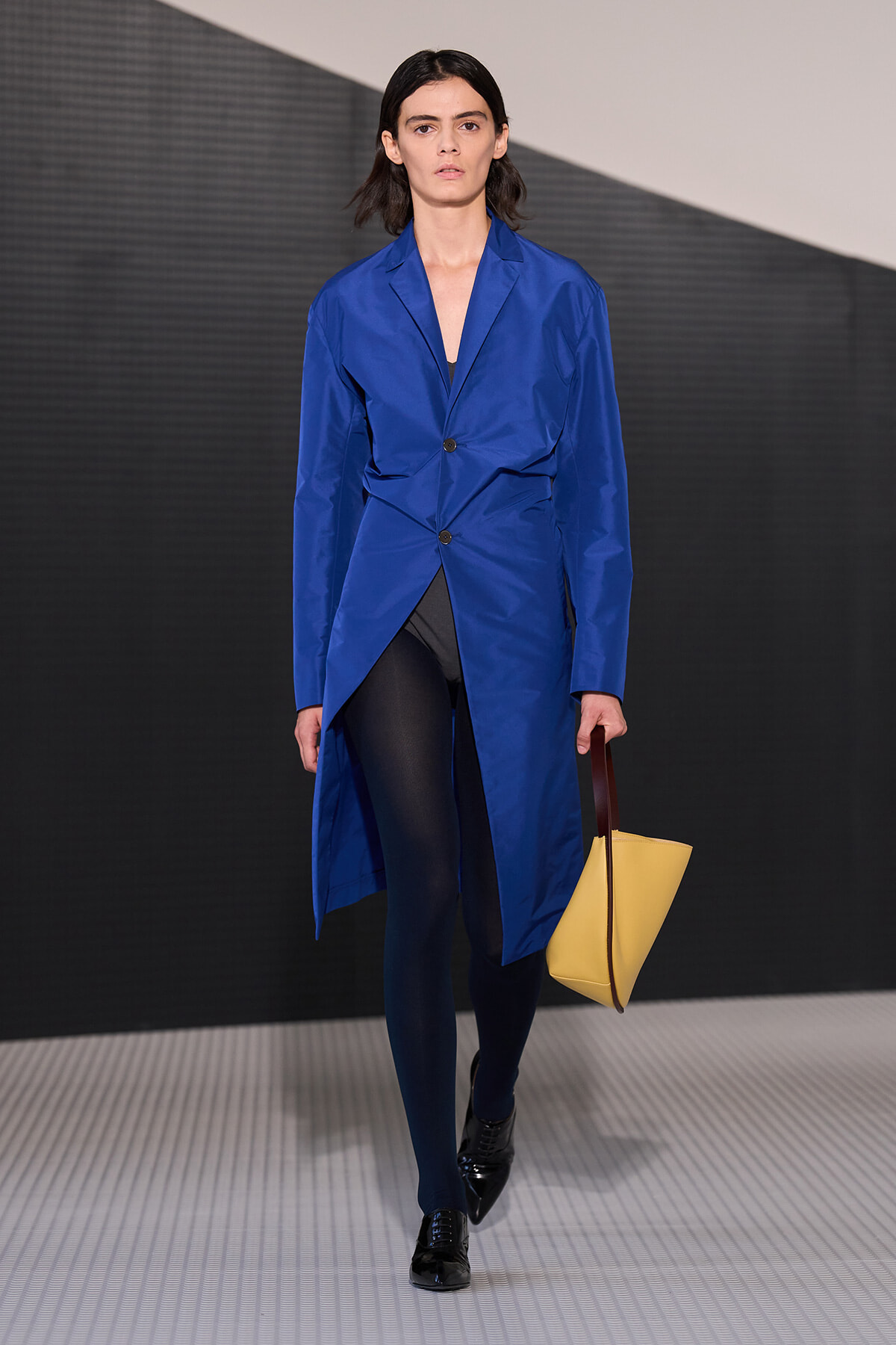 Model walks the runway in a bright blue overcoat-style blazer with an asymmetric wrap, black tights, black shiny shoes, and a yellow tote with brown handles.