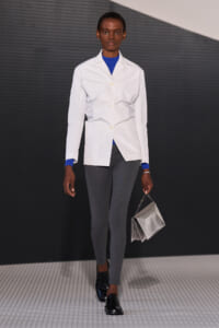 Model walking a runway in a white blazer over a blue top, gray slim pants, black shoes, and a metallic silver handbag.