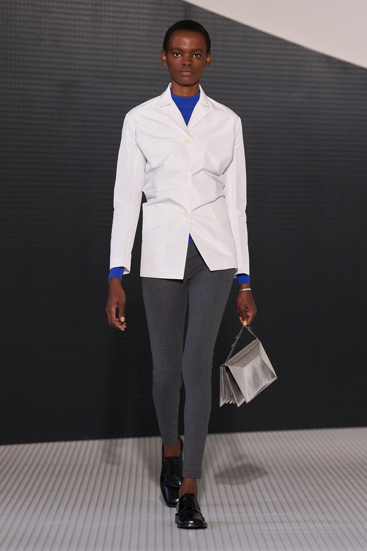 Model walking a runway in a white blazer over a blue top, gray slim pants, black shoes, and a metallic silver handbag.