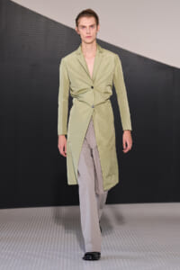 Male model walks a runway in a pale olive-green, knee-length trench with a double-breasted, wrap-style front and high front slit, paired with light taupe trousers and black shoes.